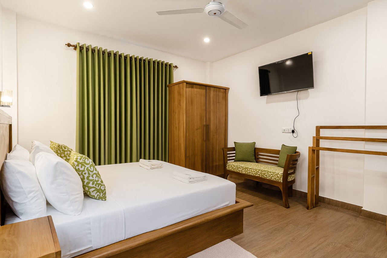 Wooden bed with green pillows and curtains, wardrobe, TV, bench, ceiling fan.