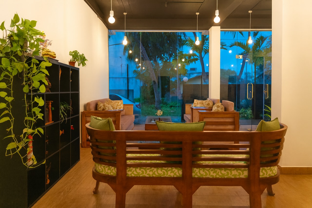 Wooden bench with green cushions, plants, large windows to palms at dusk, string lights.