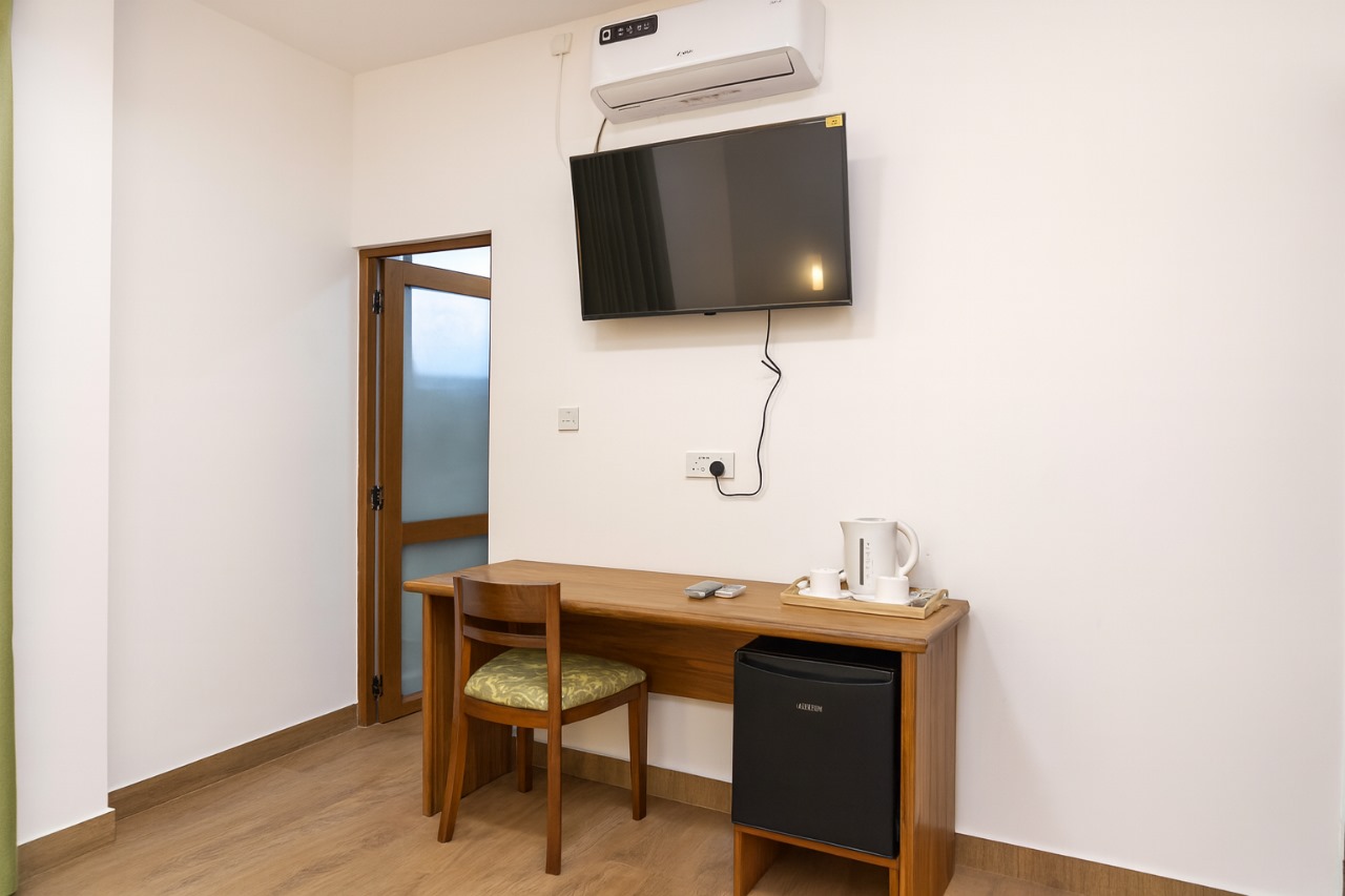 Wooden desk with green chair, mini-fridge, smart TV, AC, kettle and cups.